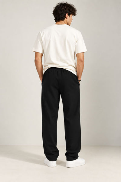 A man viewed from behind wearing a white t-shirt, black sweatpants, and white sneakers, standing against a neutral light gray studio background.