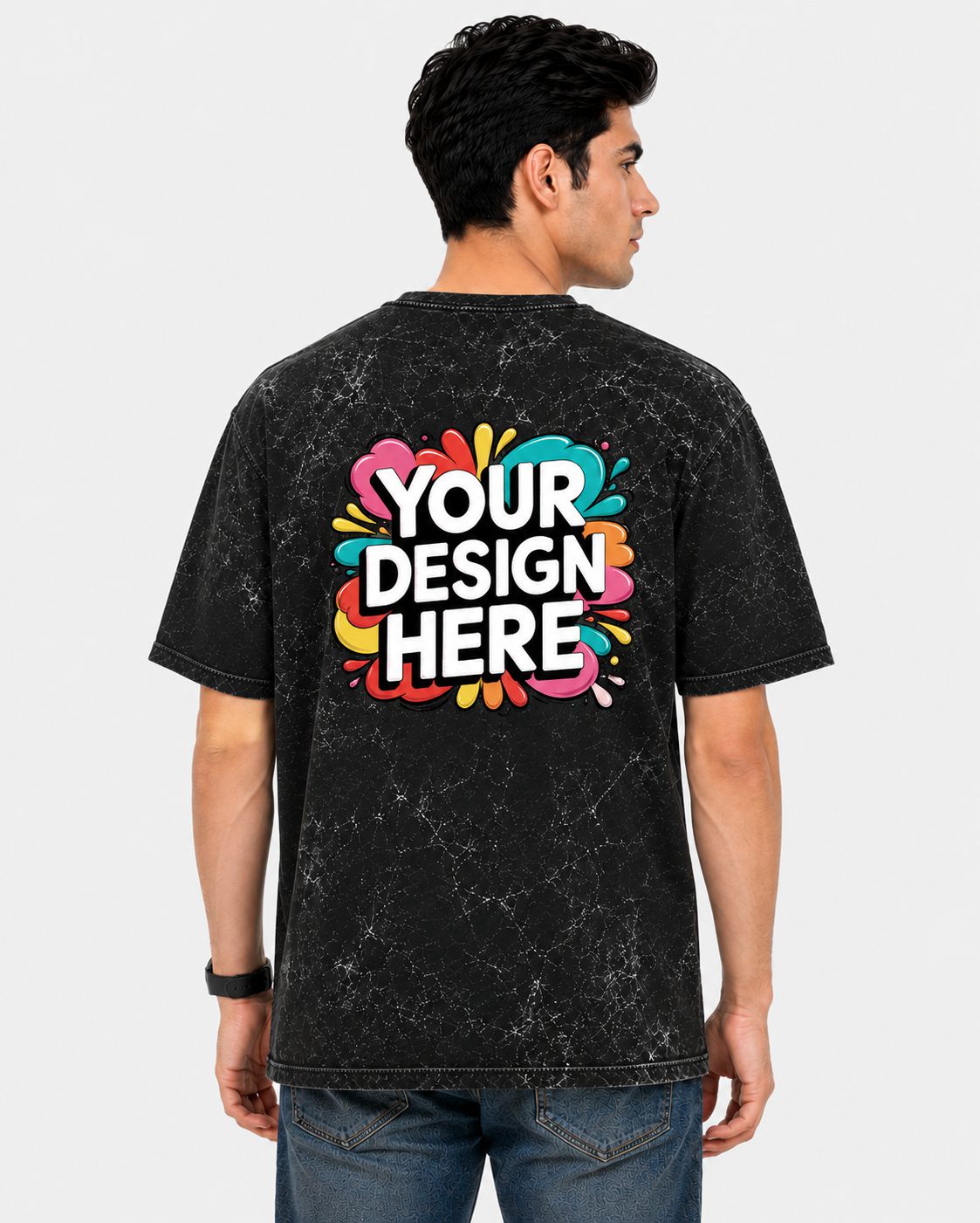 A man viewed from the back wearing a black acid-wash t-shirt with a colorful 'YOUR DESIGN HERE' mockup graphic on the back, paired with blue jeans against a light gray background.