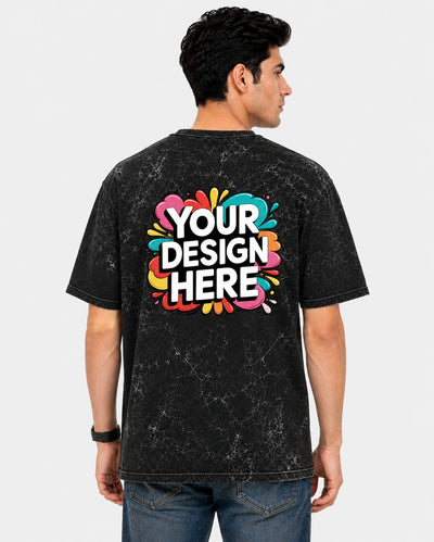 A man viewed from the back wearing a black acid-wash t-shirt with a colorful 'YOUR DESIGN HERE' mockup graphic on the back, paired with blue jeans against a light gray background.