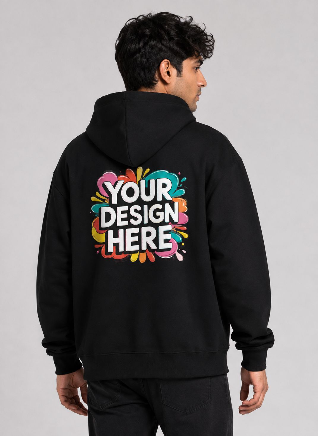 A man viewed from the back wearing a black hoodie with a colorful 'YOUR DESIGN HERE' mockup graphic printed on the back, set against a plain light gray studio background.