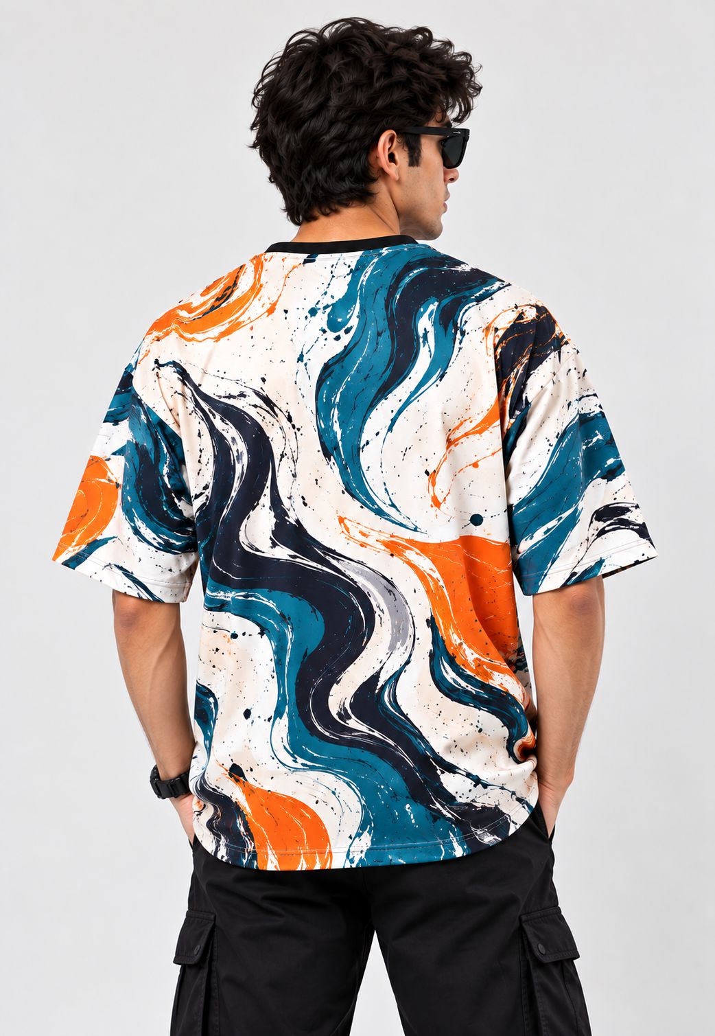 A man viewed from the back wearing a short-sleeve crewneck t-shirt with a bold abstract swirl pattern in orange, teal blue, black, and white, paired with black cargo pants against a light gray background.