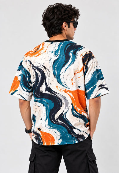 A man viewed from the back wearing a short-sleeve crewneck t-shirt with a bold abstract swirl pattern in orange, teal blue, black, and white, paired with black cargo pants against a light gray background.
