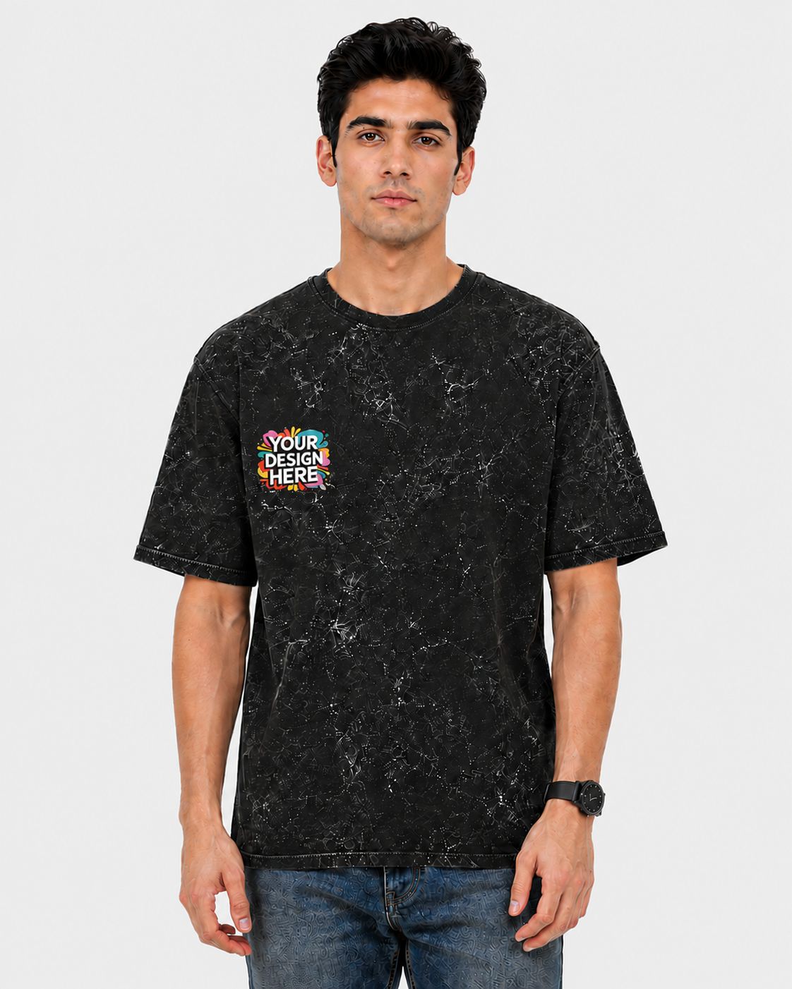 A man wearing a black mineral-wash t-shirt with a colorful 'YOUR DESIGN HERE!' graphic on the left chest, paired with blue jeans, against a light gray background.