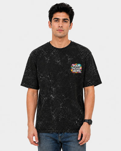 A man wearing a black mineral-wash t-shirt with a colorful 'YOUR DESIGN HERE' placeholder graphic on the left chest, paired with blue jeans, against a light gray background.