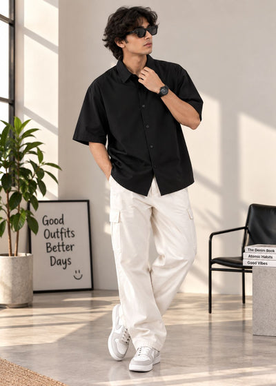 A man wearing a black short-sleeve button-up shirt, off-white cargo pants, and white sneakers, standing in a bright, modern room with a plant and framed art in the background.