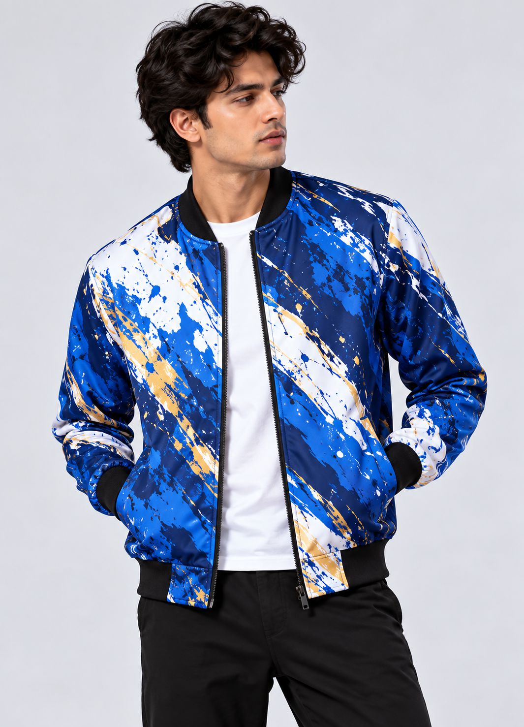 A man wearing a blue, white, and gold abstract patterned bomber jacket over a white t-shirt and black pants, photographed against a light gray studio background.