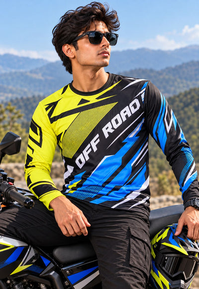 A man wearing a bright yellow, black, and blue long-sleeve off-road jersey sits on a motorcycle outdoors with mountains in the background. The jersey has 'OFF ROAD' printed across the chest.