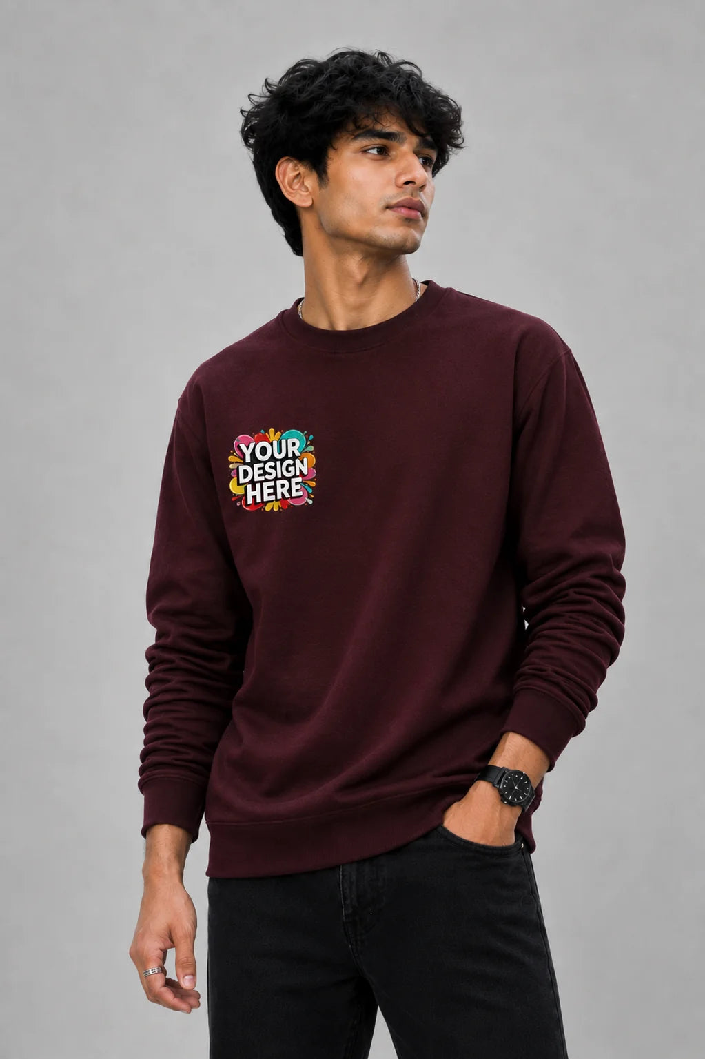 A man wearing a burgundy crewneck sweatshirt with a colorful 'YOUR DESIGN HERE' mockup graphic on the left chest, paired with black pants, standing against a neutral gray studio background.