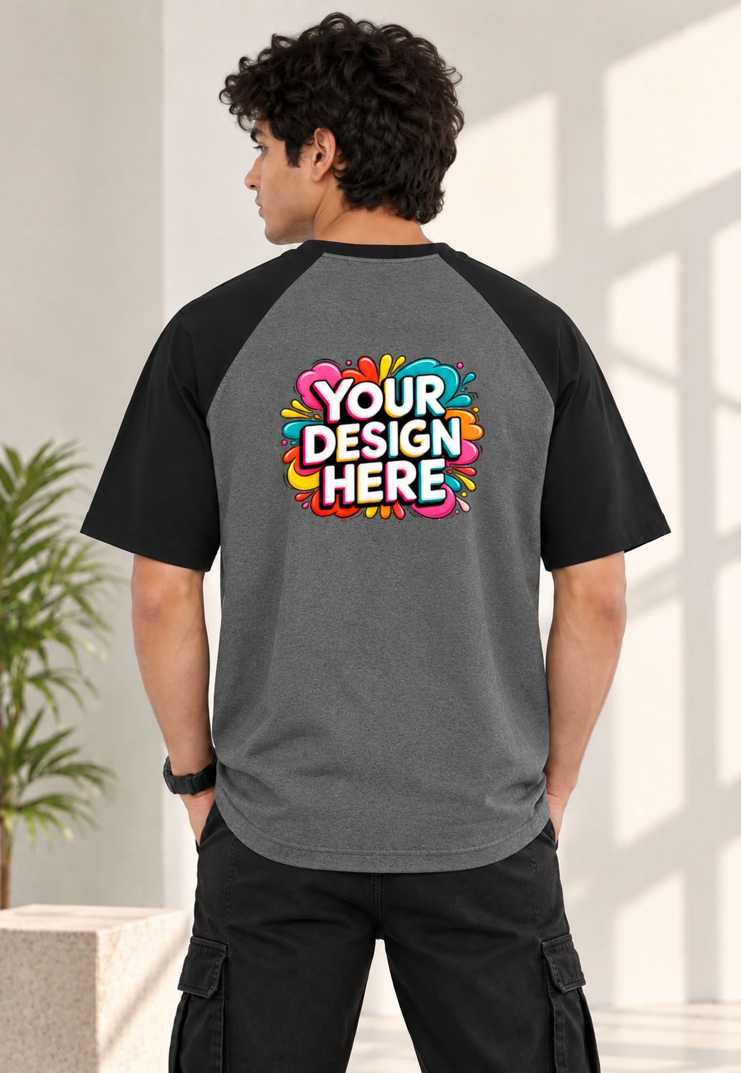 A man wearing a dark gray and black raglan baseball t-shirt from the back, featuring a colorful 'YOUR DESIGN HERE' mockup graphic on the back. The setting is a bright, modern indoor space with soft window light.