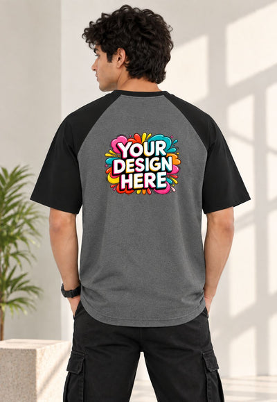 A man wearing a dark gray and black raglan baseball t-shirt from the back, featuring a colorful 'YOUR DESIGN HERE' mockup graphic on the back. The setting is a bright, modern indoor space with soft window light.