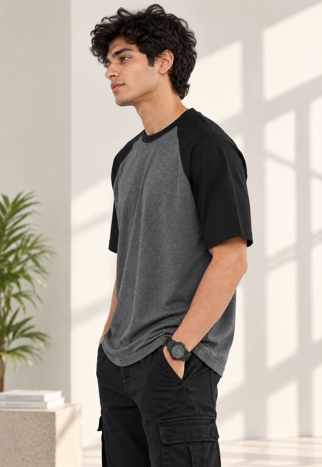 A man wearing a dark gray and black raglan t-shirt, black cargo pants, and a black watch, standing in a brightly lit room with window shadows.