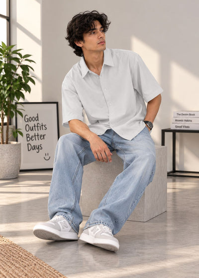 A man wearing a light gray short-sleeve button-up shirt, light blue jeans, and white sneakers sits on a concrete block in a bright, modern room.