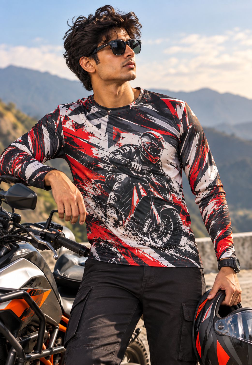 A man wearing a long-sleeve graphic jersey with a red, black, and white motorcycle racer print, standing next to a motorcycle outdoors with mountains in the background.