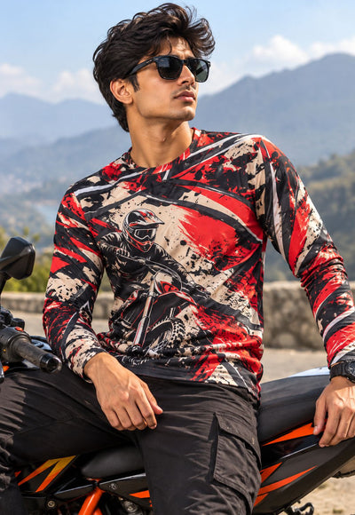 A man wearing a long-sleeve motocross jersey with a red, black, and beige graphic print sits on an orange and black motorcycle outdoors with mountains in the background.