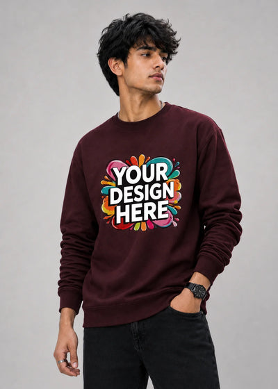 A man wearing a maroon crewneck sweatshirt with a colorful 'YOUR DESIGN HERE' mockup graphic on the chest, paired with black jeans, against a plain gray studio background.