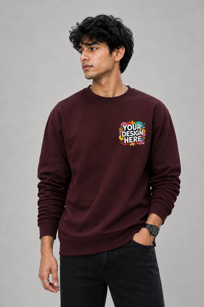 A man wearing a maroon crewneck sweatshirt with a colorful 'YOUR DESIGN HERE' mockup graphic on the left chest, paired with black jeans, against a solid gray studio background.