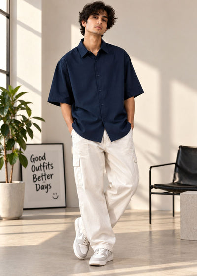 A man wearing a navy blue short-sleeve button-up shirt and off-white cargo pants, standing in a bright, modern room with a potted plant and framed art in the background.