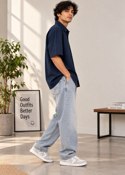 A man wearing a navy blue short-sleeve button-up shirt, light-wash wide-leg jeans, and white sneakers, standing in a bright, modern room with a plant and framed art in the background.