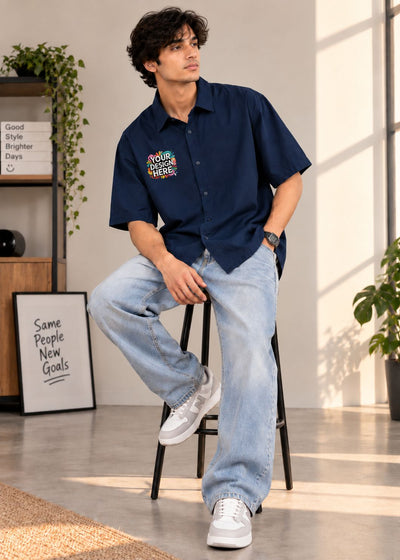 A man wearing a navy blue short-sleeve button-up shirt with a colorful 'YOUR DESIGN HERE' graphic on the left chest, paired with light-wash jeans and white sneakers, sitting on a black stool in a bright room.