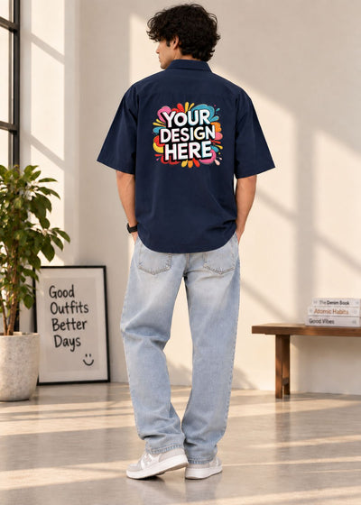 A man wearing a navy blue short-sleeve button-up shirt with a colorful 'YOUR DESIGN HERE' graphic printed on the back, paired with light-wash jeans and white sneakers, standing in a bright indoor room.