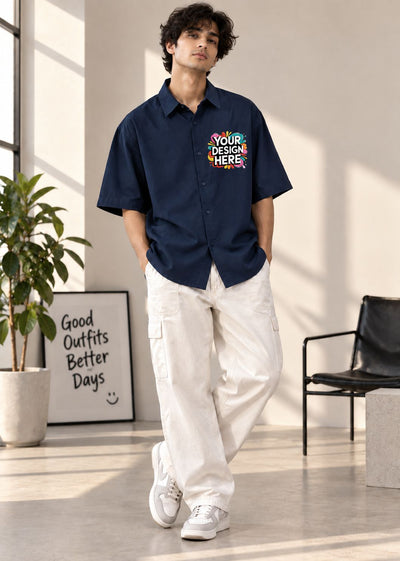 A man wearing a navy blue short-sleeve button-up shirt with a colorful 'YOUR DESIGN HERE' mockup graphic on the left chest, paired with off-white cargo pants and white sneakers, standing in a bright, modern room.
