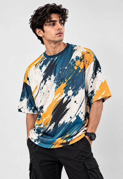 A man wearing a short-sleeve crewneck t-shirt with an abstract paint splatter pattern in blue, yellow, black, and off-white, paired with black cargo pants against a light gray studio background.