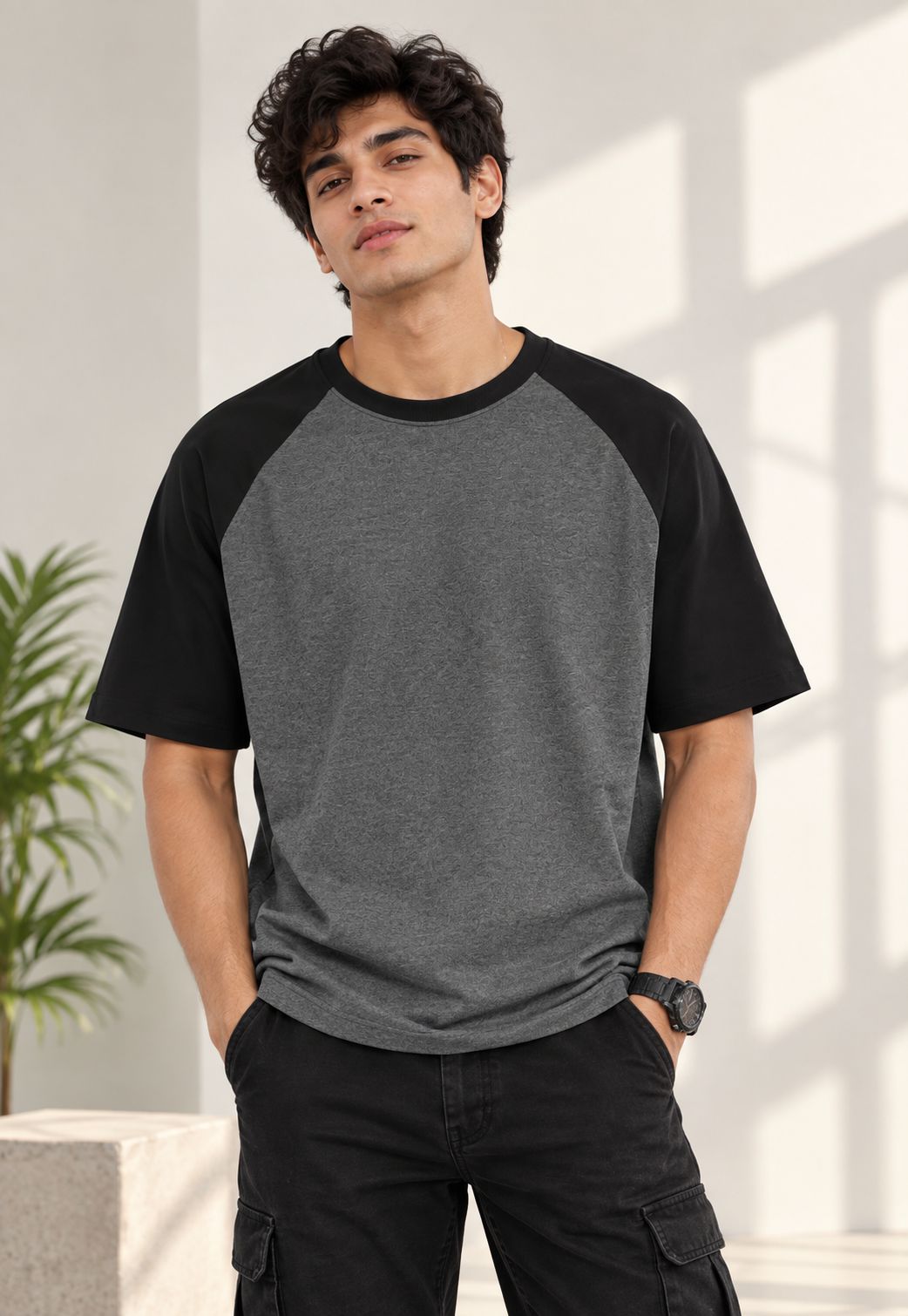 A man wearing a short-sleeve raglan t-shirt with a heather gray body and black sleeves, paired with black cargo pants, standing in a brightly lit indoor setting.