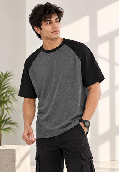 A man wearing a short-sleeve raglan t-shirt with a heather gray body and black sleeves, paired with black cargo pants, standing in a brightly lit room with window shadows.
