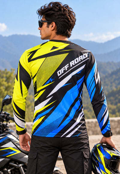 A man wearing a vibrant yellow, black, and blue long-sleeve motocross jersey with 'OFF ROAD' printed on the back, standing next to a motorcycle with a mountain background.
