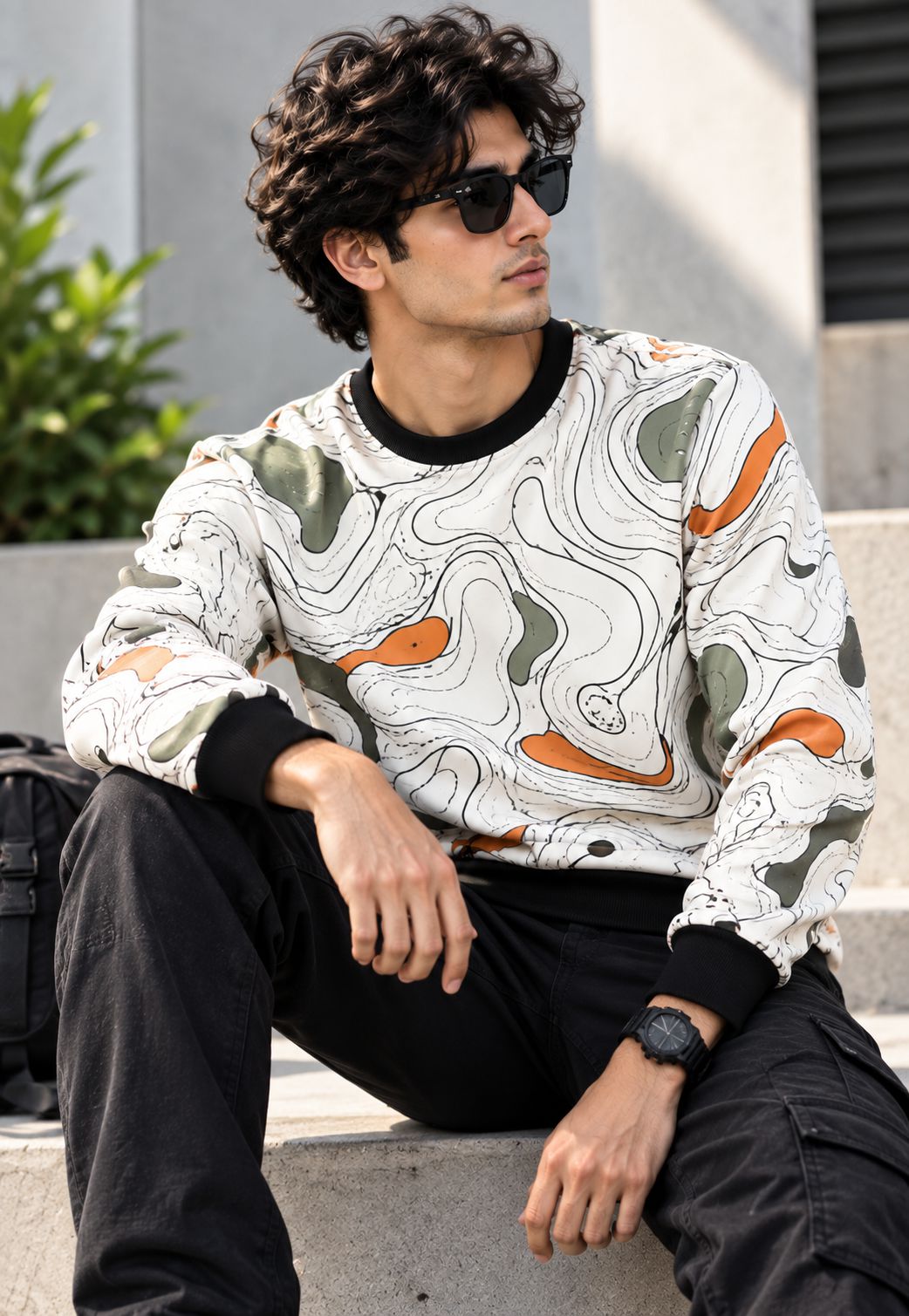 A man wearing a white long-sleeve crewneck sweatshirt with an abstract wavy pattern in orange, olive green, and gray, paired with black pants and sunglasses, sitting on outdoor concrete steps.