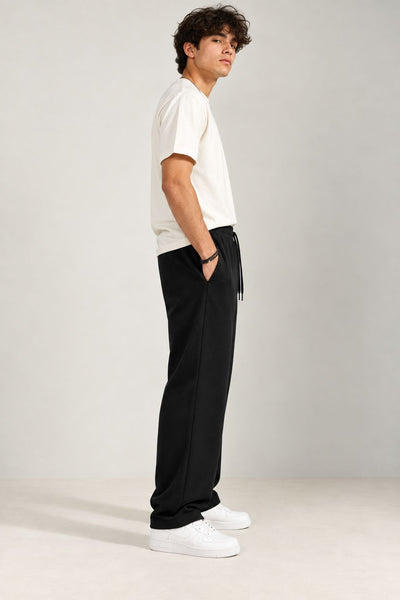 A man wearing a white t-shirt, black drawstring sweatpants, and white sneakers, standing in profile against a light gray studio background.