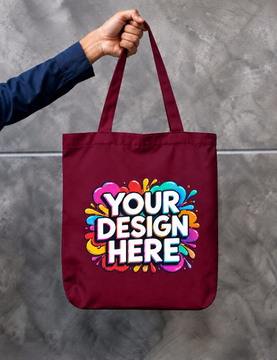 A maroon canvas tote bag held by a person's hand against a gray concrete wall. The bag features a colorful graphic with the text 'YOUR DESIGN HERE' in white with a black outline, surrounded by vibrant paint splatters.