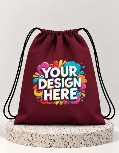 A maroon drawstring bag with black cords, featuring a colorful 'YOUR DESIGN HERE' mockup graphic on the front, displayed on a round speckled stone pedestal against a light gray background.