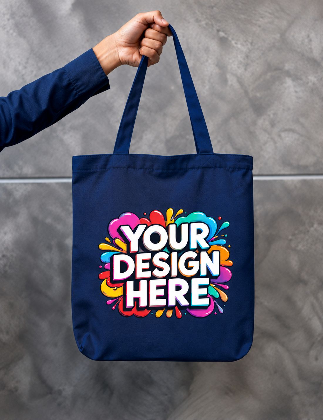 A navy blue canvas tote bag held by a person's hand against a gray textured wall. The bag features a colorful paint splatter graphic with the text 'YOUR DESIGN HERE' in white with a black outline.