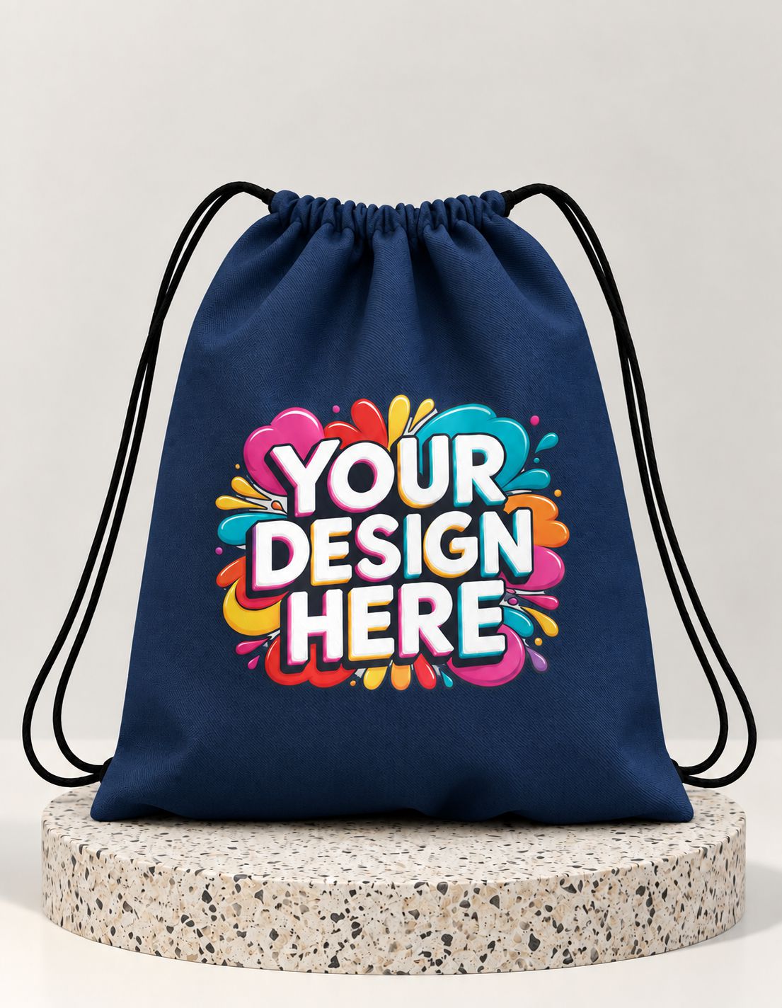 A navy blue drawstring bag with black cords, featuring a colorful 'YOUR DESIGN HERE' graphic on the front, displayed on a round speckled stone pedestal against a light gray background.