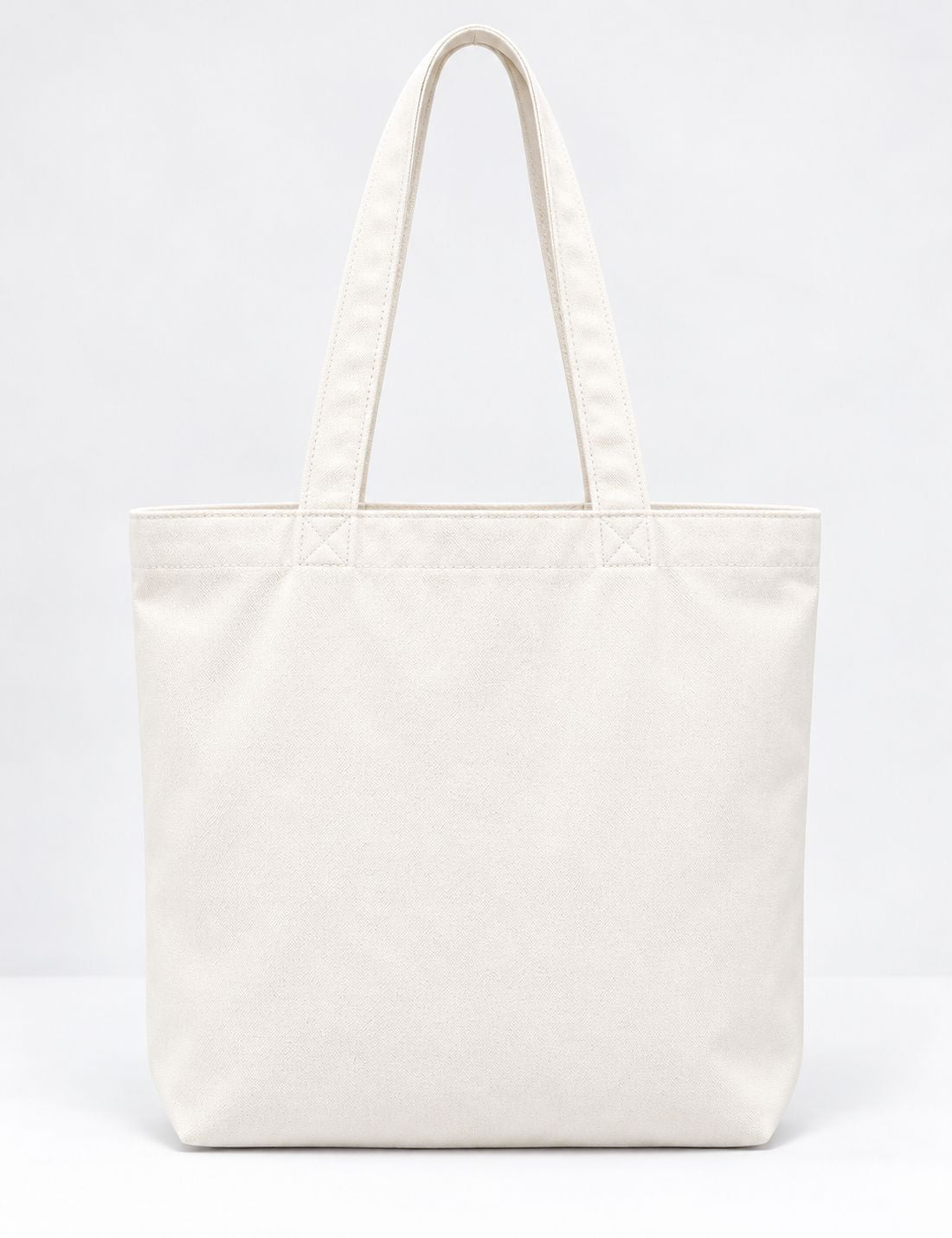 A plain canvas tote bag in off-white with dual shoulder straps, photographed against a seamless white studio background.