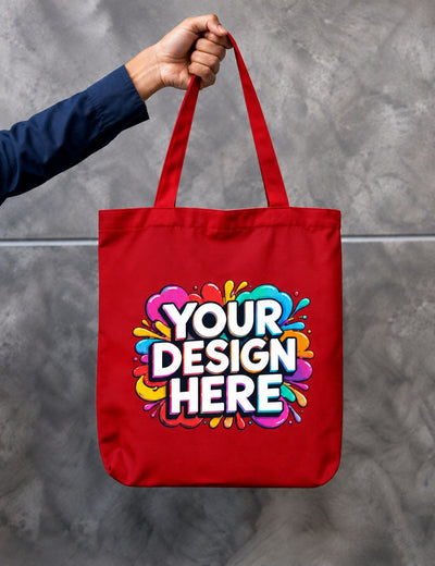 A red canvas tote bag held by a person's hand against a gray concrete wall. The bag features a colorful paint splatter graphic with the text 'YOUR DESIGN HERE' in white with a black outline.