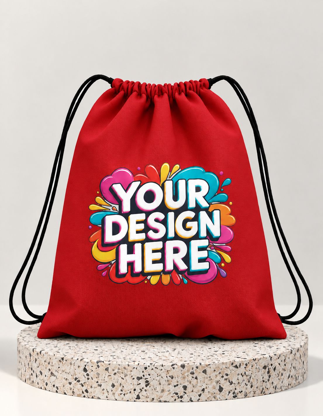 A red drawstring bag with black cords, featuring a colorful 'YOUR DESIGN HERE' mockup graphic on the front, displayed on a round speckled stone pedestal against a light gray background.