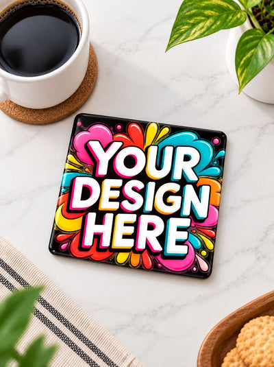 A square black coaster with a colorful graffiti-style design featuring the text 'YOUR DESIGN HERE' in bold white letters, styled on a white marble tabletop with a cup of coffee, a plant, and a striped napkin nearby.