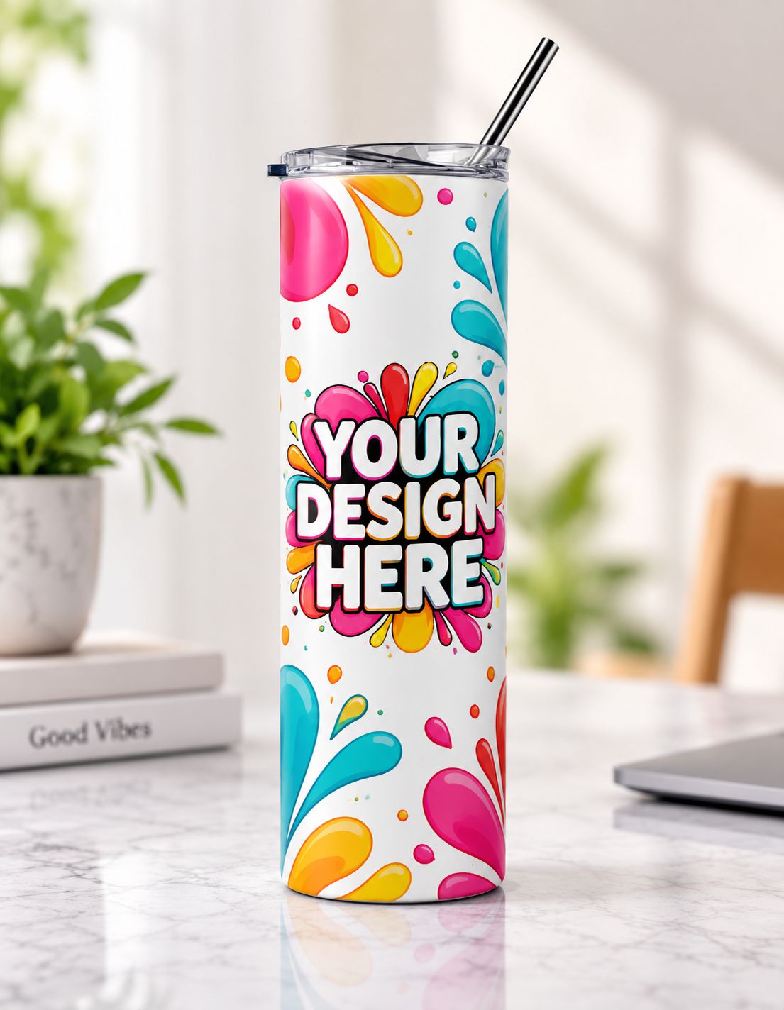 A tall insulated tumbler with a clear lid and metal straw stands on a marble table. The tumbler has a white base with colorful paint splashes in pink, blue, yellow, and orange, and the words 'YOUR DESIGN HERE' printed on the front.