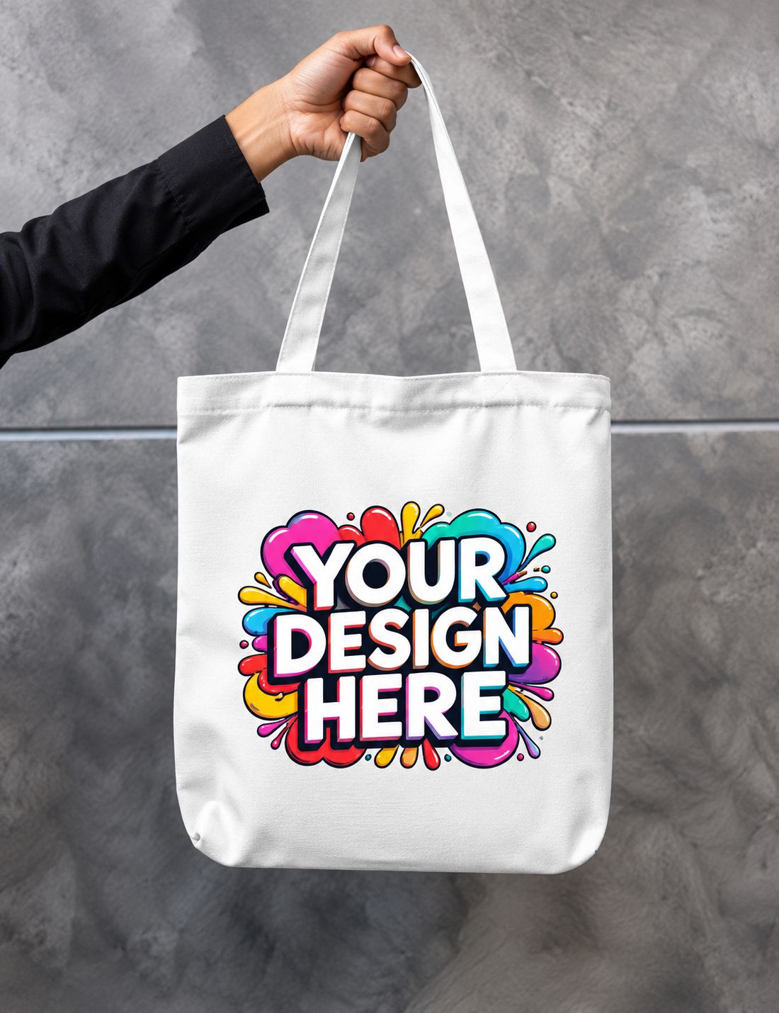 A white canvas tote bag held by a person's hand against a gray textured wall. The bag features a colorful paint-splatter graphic with the text 'YOUR DESIGN HERE' in bold white letters with a black outline.