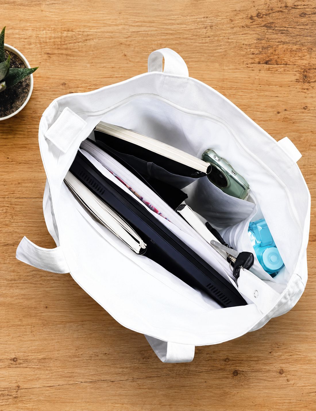 A white canvas tote bag shown open from above on a wooden tabletop, filled with notebooks, a tablet, pens, a small bottle, and other office accessories.