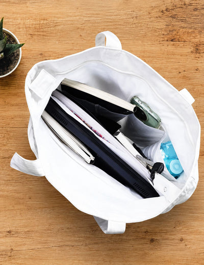 A white canvas tote bag shown open from above on a wooden tabletop, filled with notebooks, a tablet, pens, a small bottle, and other office accessories.