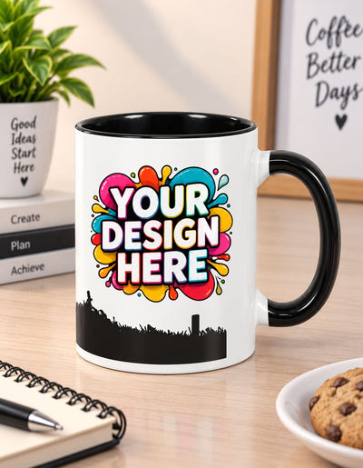 A white ceramic coffee mug with a black handle and interior, featuring a colorful 'YOUR DESIGN HERE' graphic and a black skyline silhouette on the front, displayed on a wooden desk with a notebook, cookie, and plant in the background.