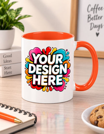 A white ceramic coffee mug with a bright orange handle and interior, featuring a colorful 'YOUR DESIGN HERE' mockup graphic on the front. The mug is set on a wooden desk with a notebook, plant, and cookies in a bright, modern room.
