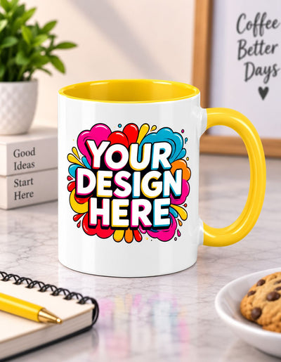 A white ceramic coffee mug with a bright yellow handle and interior, featuring a colorful abstract heart design with the text 'YOUR DESIGN HERE' in white. The mug is set on a marble desk with a notebook, plant, and cookie in a lifestyle setting.