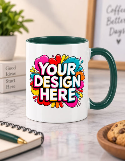A white ceramic coffee mug with a dark green handle and interior, featuring a colorful 'YOUR DESIGN HERE' placeholder graphic on the front, set on a marble desk with a notebook, plant, and cookie in the background.