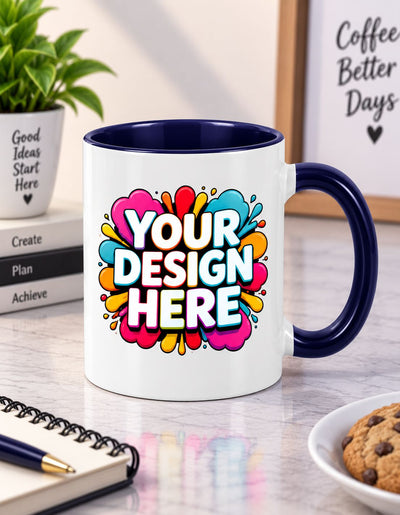 A white ceramic coffee mug with a navy blue handle and interior, featuring a colorful 'YOUR DESIGN HERE' placeholder graphic on the front. The mug is set on a marble desk with a notebook, cookie, and a potted plant in the background.
