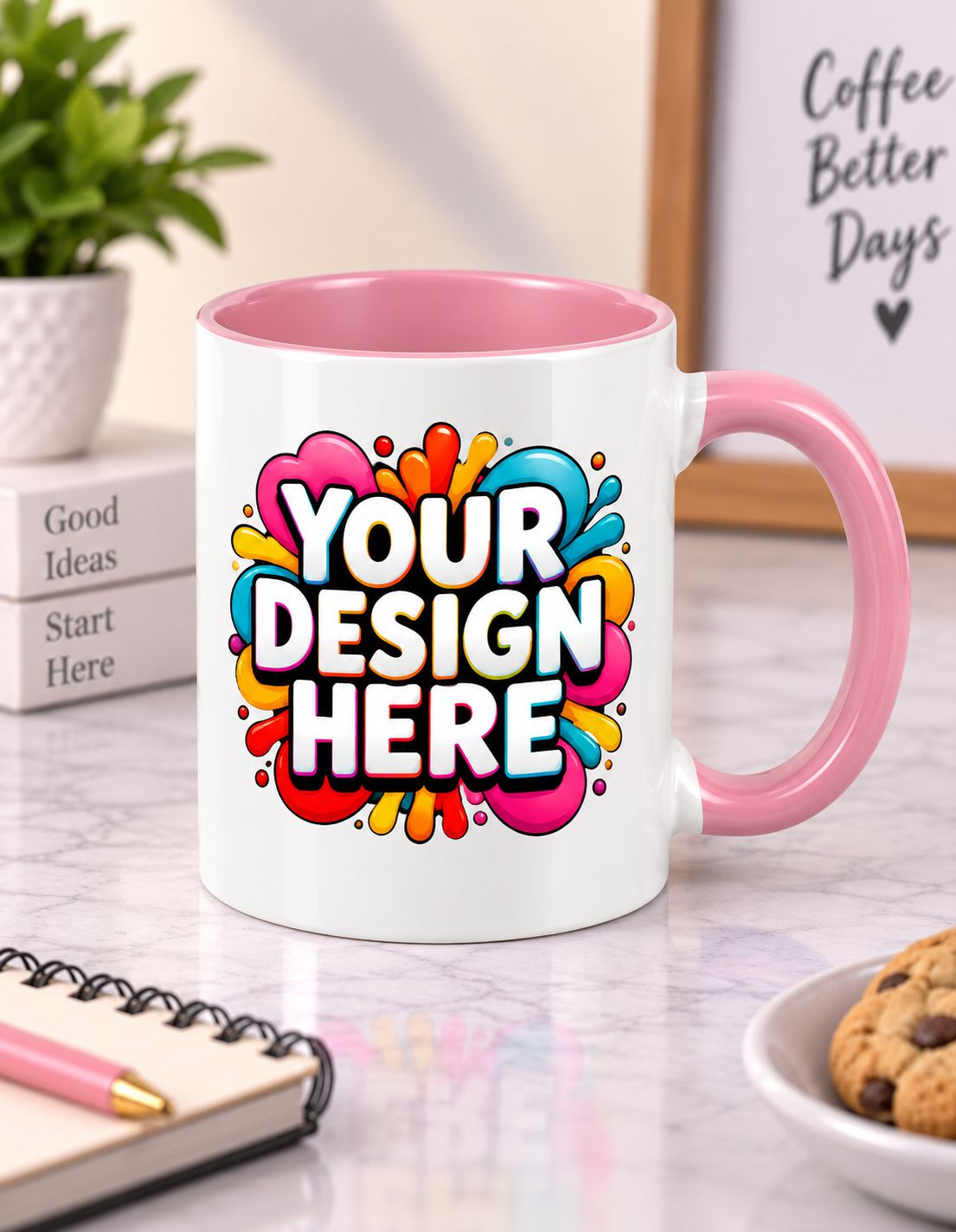 A white ceramic coffee mug with a pink handle and interior, featuring a colorful paint splatter graphic with the text 'YOUR DESIGN HERE'. The mug is set on a marble desk with a notebook, a cookie, and a plant in the background.