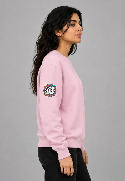 A woman in a light pink crewneck sweatshirt with a customizable circular patch on the left sleeve, standing against a solid gray background.